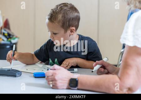 Ein kleiner Junge sitzt mit einem Lehrer an einem Tisch und schreibt mit Markierungen. Der Lehrer beobachtet den Jungen und scheint ihm bei seiner Arbeit zu helfen. Die Stockfoto