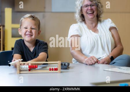 Ein kleiner Junge sitzt mit einem Abakus an einem Tisch und lächelt mit seinem Lehrer, der mit ihm lacht, als er das Problem löst. Stockfoto