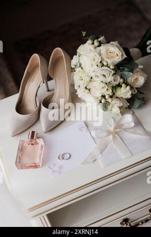 Brautjungfernkostüme: Absatzschuhe, Blumenstrauß, Ringe, Parfüm und Einladung auf einem weißen Tisch Stockfoto