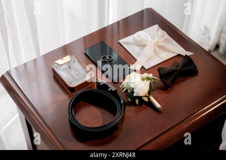 Die wichtigsten Hochzeitsartikel des Bräutigams auf dem Tisch, darunter Ringe am Telefon, Fliege, Boutonniere, Parfüm und Gürtel Stockfoto