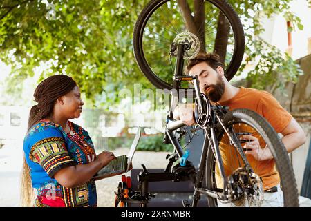 Begeistertes, sportliches multiethnisches Paar mit Laptop, das Fahrrad im Hof repariert. Gesunder, engagierter kaukasischer Mann und afroamerikanische Frau, die auf Laptop suchen, um kaputte Fahrradketten und Pedale zu reparieren. Stockfoto