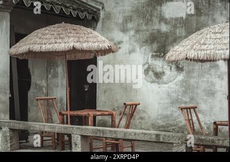 Vor einer Betonwand befindet sich eine Terrasse mit Bambusmöbeln und Strohschirmen. Stockfoto