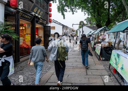 Suzhou, China - 11. Juni 2024 : Eine enge Kopfsteinpflasterstraße in Suzhou, China, gesäumt von traditionellen Geschäften und Marktständen. Stockfoto