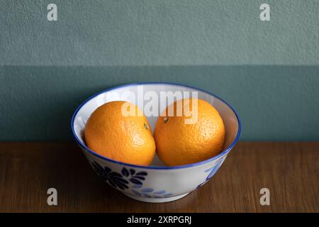 Zwei Orangen Obst auf einer weißen Keramikschale auf dem braunen Teaktisch, dunkelgrüne Wand im Hintergrund. Stockfoto