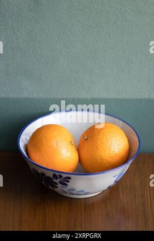 Zwei Orangen Obst auf einer weißen Keramikschale auf dem braunen Teaktisch, dunkelgrüne Wand im Hintergrund. Stockfoto