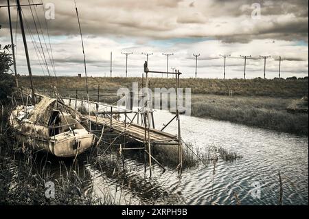 Altes Segelboot neben dem maroden Startsteg auf dem Fluss bei Freckleton, Lancashire, mit Landebahn-Landebampen im Hintergrund Stockfoto