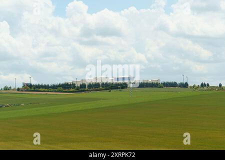 Ein üppiges, grünes Feld erstreckt sich unter einem hellblauen Himmel mit flauschigen weißen Wolken. In der Ferne ein großer Industriekomplex mit weißem bui Stockfoto