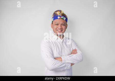 Porträt eines Balinesen mit traditionellem Kopfschmuck namens Udeng, der mit überkreuzten Armen und selbstbewusstem Lächeln über isoliertem weißen Hintergrund posiert Stockfoto