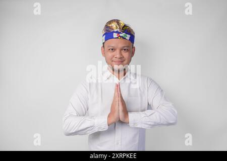 Lächelnder junger balinesischer Mann mit traditionellem Kopfschmuck namens Udeng, der Begrüßungs- oder Willkommensgesten tut, isoliert über weißem Hintergrund Stockfoto