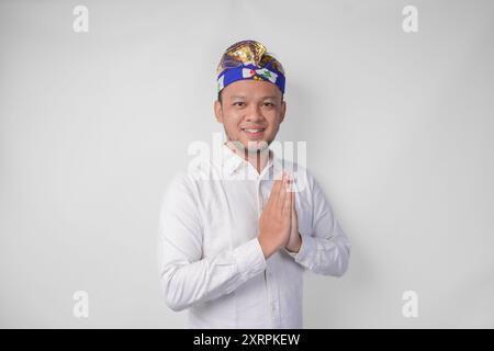 Lächelnder junger balinesischer Mann mit traditionellem Kopfschmuck namens Udeng, der Begrüßungs- oder Willkommensgesten tut, isoliert über weißem Hintergrund Stockfoto