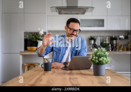 Fröhlicher junger männlicher Profi, der die Hand winkt, während Videokonferenzen über Laptop im Home Office Stockfoto