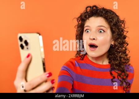 Eine schockierte junge Frau mit lockigen Haaren starrt auf ihr Handy und trägt einen gestreiften Pullover vor orangem Hintergrund. Stockfoto