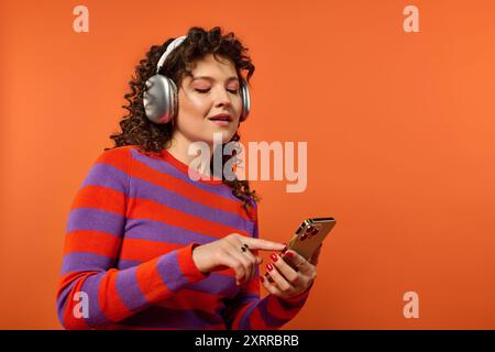 Eine junge Frau mit lockigen Haaren posiert in einem farbenfrohen gestreiften Pullover vor einem leuchtend orangefarbenen Hintergrund. Stockfoto