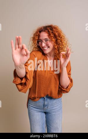 Aufgeregte Freelancerin mit Rothaarigen, die OK-Geste zeigt, während sie auf dem Hintergrund steht Stockfoto