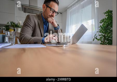 Fokussierter junger Geschäftsmann im Anzug, der das Kinn berührt und im Heimbüro auf dem Schreibtisch sitzt Stockfoto