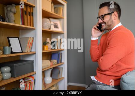 Seitenansicht eines jungen Geschäftsmannes, der über das Smartphone spricht, während er zu Hause vor dem Bücherregal sitzt Stockfoto