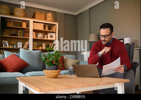 Fokussierter Geschäftsmann mit Dokumenten, die Informationen online über ein Notebook auf dem Tisch im Heimbüro lesen Stockfoto