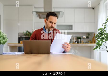 Fokussierter junger Unternehmer, der Geschäftsberichte analysiert und im Home Office am Schreibtisch über ein Notebook arbeitet Stockfoto