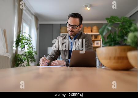 Fokussierter junger, eleganter Geschäftsmann, der Dokumente unterschreibt, während er am Schreibtisch im Heimbüro am Laptop arbeitet Stockfoto