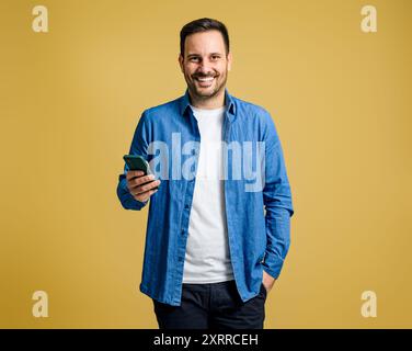 Lächelnder, gutaussehender junger Mann, der auf dem Handy in sozialen Medien surft, während er auf gelbem Hintergrund steht Stockfoto