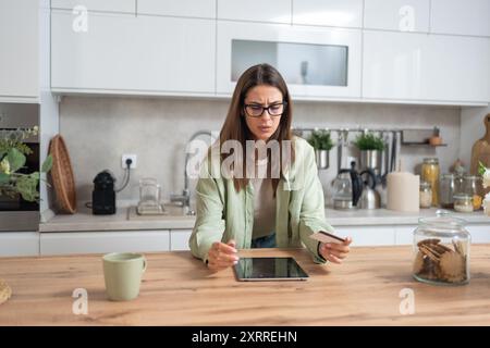 Junge verwirrte Frau mit Kreditkarte, die auf dem Bildschirm eines digitalen Tablets schaut, Probleme mit Online-Banking oder -Shopping, Kreditkartenlimit und Überspende hat Stockfoto