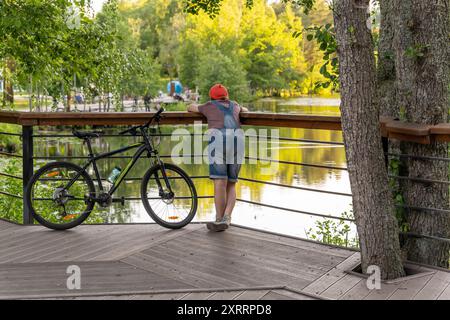 Wooman im Overall lehnt sich auf das Geländer, genießt den ruhigen Blick auf den See, Fahrrad Stockfoto