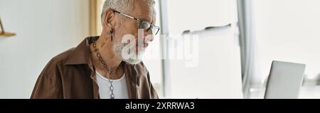 Ein reifer schwuler Mann mit Tattoos und grauen Haaren arbeitet zu Hause ferngesteuert auf seinem Laptop. Stockfoto