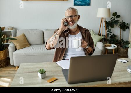 Ein reifer schwuler Mann mit Tattoos und grauen Haaren lächelt, während er an einem Arbeitstag am Telefon spricht und Kaffee trinkt. Stockfoto