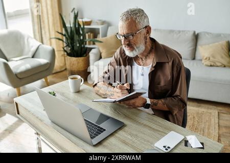 Ein reifer schwuler Mann mit Tattoos und grauen Haaren arbeitet von zu Hause aus an seinem Laptop. Stockfoto