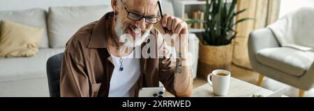 Ein reifer schwuler Mann mit Tattoos und grauen Haaren lächelt, während er von zu Hause aus arbeitet. Stockfoto