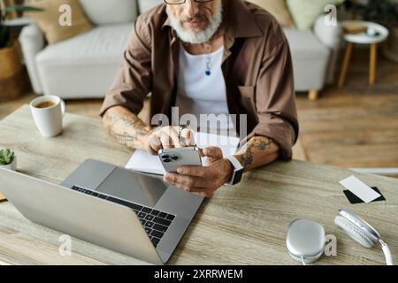 Ein reifer schwuler Mann mit grauem Haar und Tattoos arbeitet von zu Hause aus mit einem Smartphone und Laptop. Stockfoto