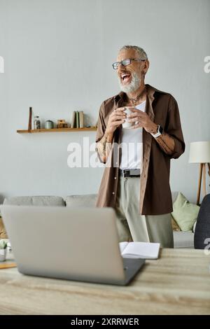 Ein reifer schwuler Mann mit Tattoos und grauem Haar lacht, während er von zu Hause aus arbeitet. Stockfoto