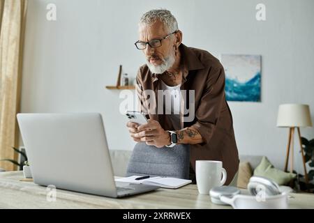 Ein reifer schwuler Mann mit Tattoos und grauen Haaren überprüft sein Handy, während er von zu Hause aus arbeitet. Stockfoto