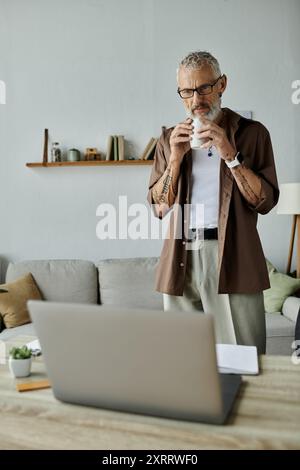 Ein reifer schwuler Mann mit Tattoos und grauem Haar, der von zu Hause aus arbeitet und eine Kaffeepause genießt. Stockfoto