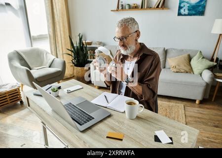 Ein reifer schwuler Mann mit Tattoos und grauen Haaren arbeitet von zu Hause aus, hält Kopfhörer und schaut sich einen Laptop an. Stockfoto