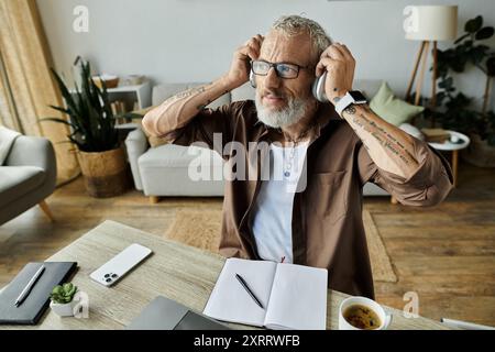 Ein reifer schwuler Mann mit Tattoos und grauen Haaren arbeitet von zu Hause aus, während er Kopfhörer trägt. Stockfoto