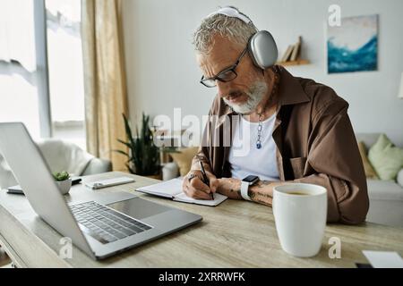 Ein reifer schwuler Mann mit Tattoos und grauen Haaren arbeitet von zu Hause aus, trägt Kopfhörer und schreibt in einem Notebook. Stockfoto