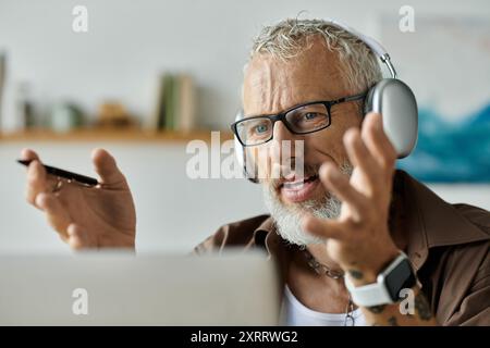 Ein reifer schwuler Mann mit grauem Haar und Tattoos arbeitet von zu Hause aus, während er Kopfhörer und Brillen trägt. Stockfoto