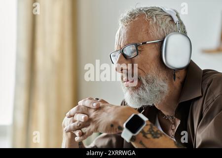 Ein reifer schwuler Mann mit Tattoos und grauem Haar arbeitet von zu Hause aus. Er trägt Brille und Kopfhörer. Stockfoto