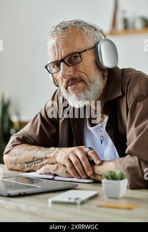 Ein reifer schwuler Mann mit Tattoos und grauen Haaren arbeitet von zu Hause aus, trägt Kopfhörer und blickt direkt in die Kamera. Stockfoto