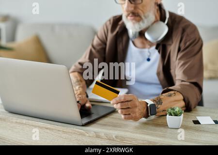 Ein reifer schwuler Mann mit Tattoos und grauen Haaren sitzt an seinem Schreibtisch, arbeitet ferngesteuert an einem Laptop und hält eine Kreditkarte. Stockfoto