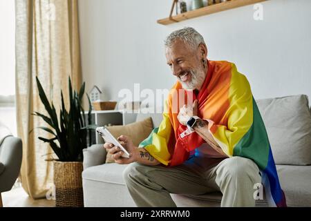 Ein reifer schwuler Mann mit grauen Haaren und Tattoos lächelt, während er eine Stolzfahne hält. Er sitzt auf einer Couch in einem Heim. Stockfoto