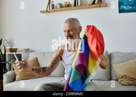 Ein reifer schwuler Mann mit Tattoos und grauen Haaren hält eine Stolz-Regenbogenfahne in seinem Wohnzimmer. Stockfoto
