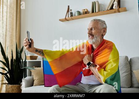 Ein reifer schwuler Mann mit Tattoos und grauen Haaren macht ein Selfie, während er zu Hause in eine Regenbogenflagge gehüllt ist. Stockfoto