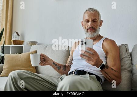 Ein reifer Mann mit Tattoos und grauem Bart entspannt sich auf einer Couch zu Hause, während er sein Telefon benutzt. Stockfoto