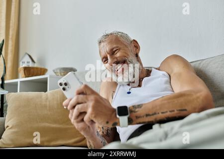 Ein tätowierter Mann mit grauem Bart entspannt sich auf einer Couch und lächelt, während er auf sein Handy schaut. Stockfoto