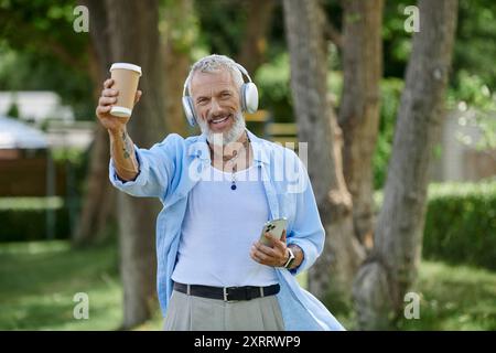 Ein älterer bärtiger Mann mit Tattoos genießt Kaffee, lächelt und hört Musik mit Kopfhörern Stockfoto