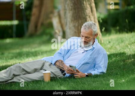 Ein Mann mit Tattoos und grauem Bart sitzt auf dem Gras und schaut auf sein Handy. Stockfoto