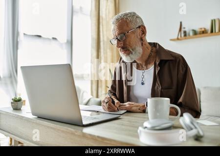 Ein reifer schwuler Mann mit Tattoos und grauen Haaren arbeitet von zu Hause aus, benutzt einen Laptop und macht Notizen. Stockfoto