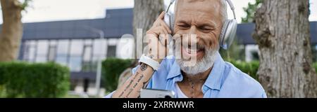 Ein reifer Mann mit grauem Bart und Tätowierungen, die hell lächeln, während sie Musik über Kopfhörer hören. Stockfoto
